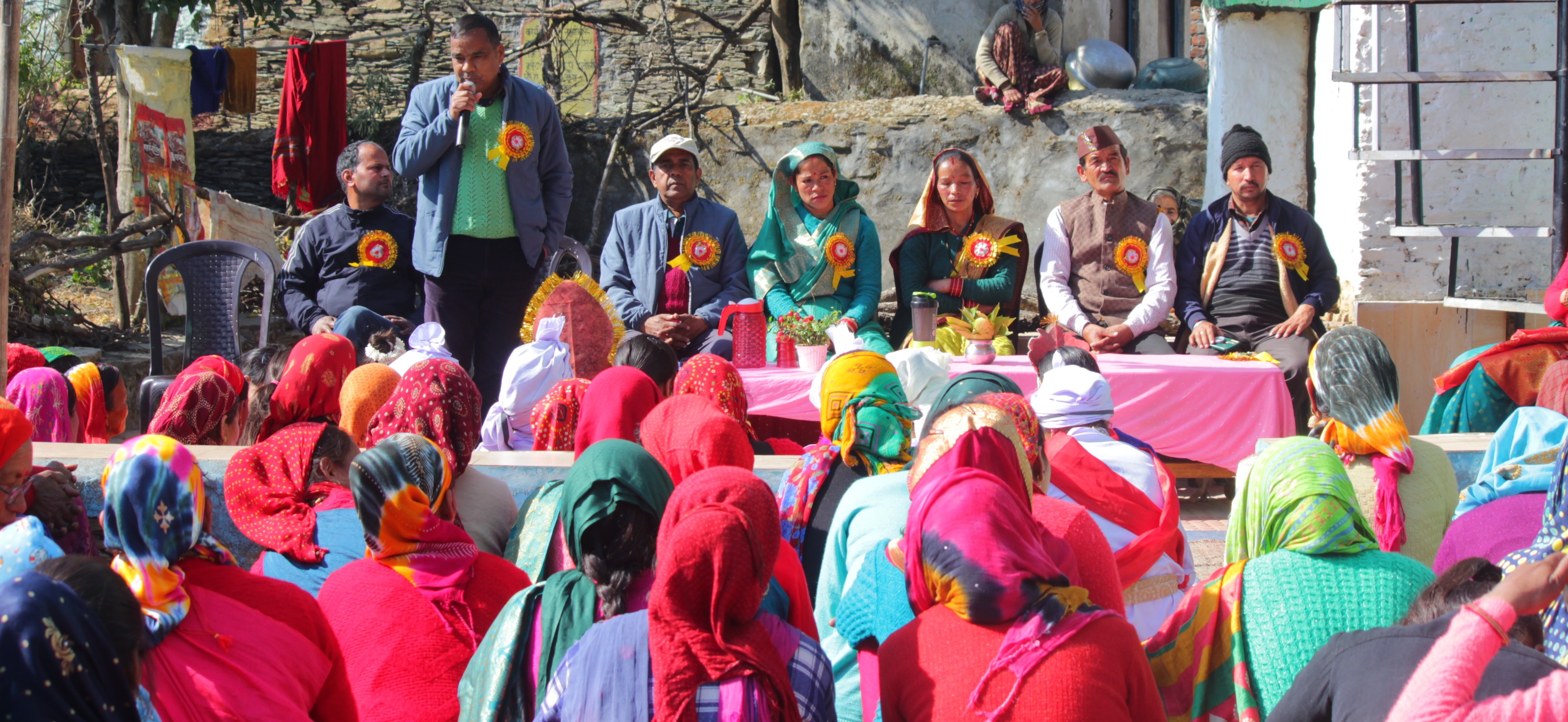 A speaker addresses an outdoor village gathering with many women in the audience.