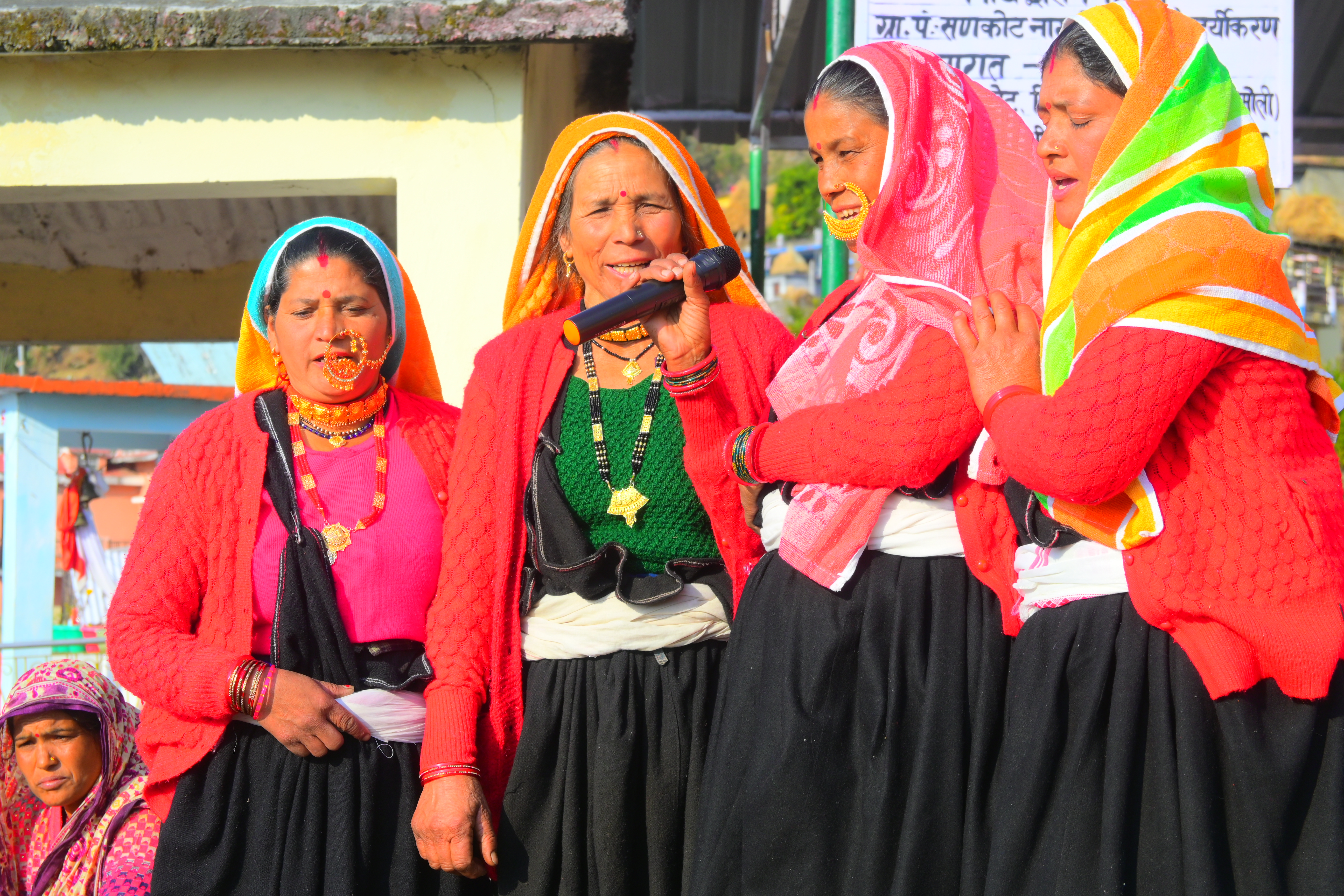 Women wearing traditional jewellery and colourful scarves sing into a microphone.
