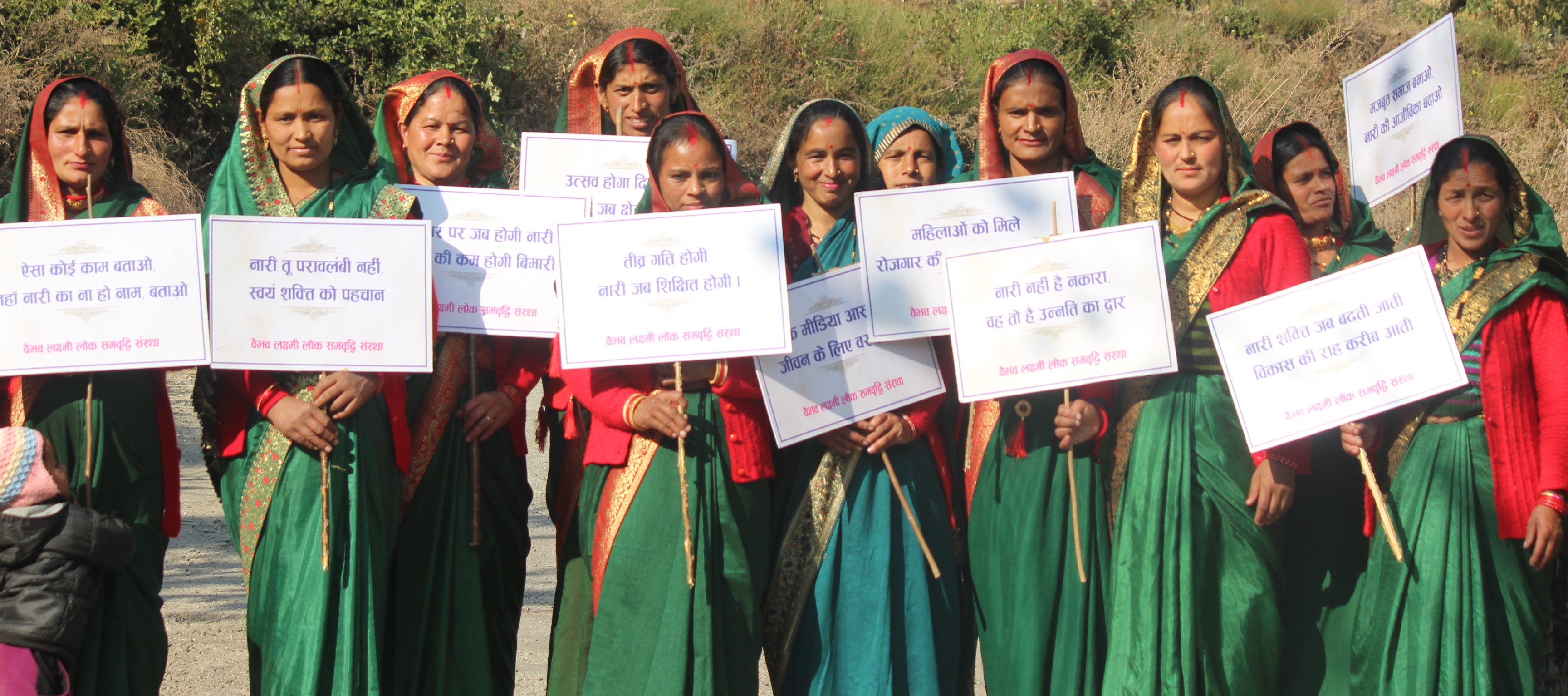 Women stand outdoors holding placards with community-awareness messages.
