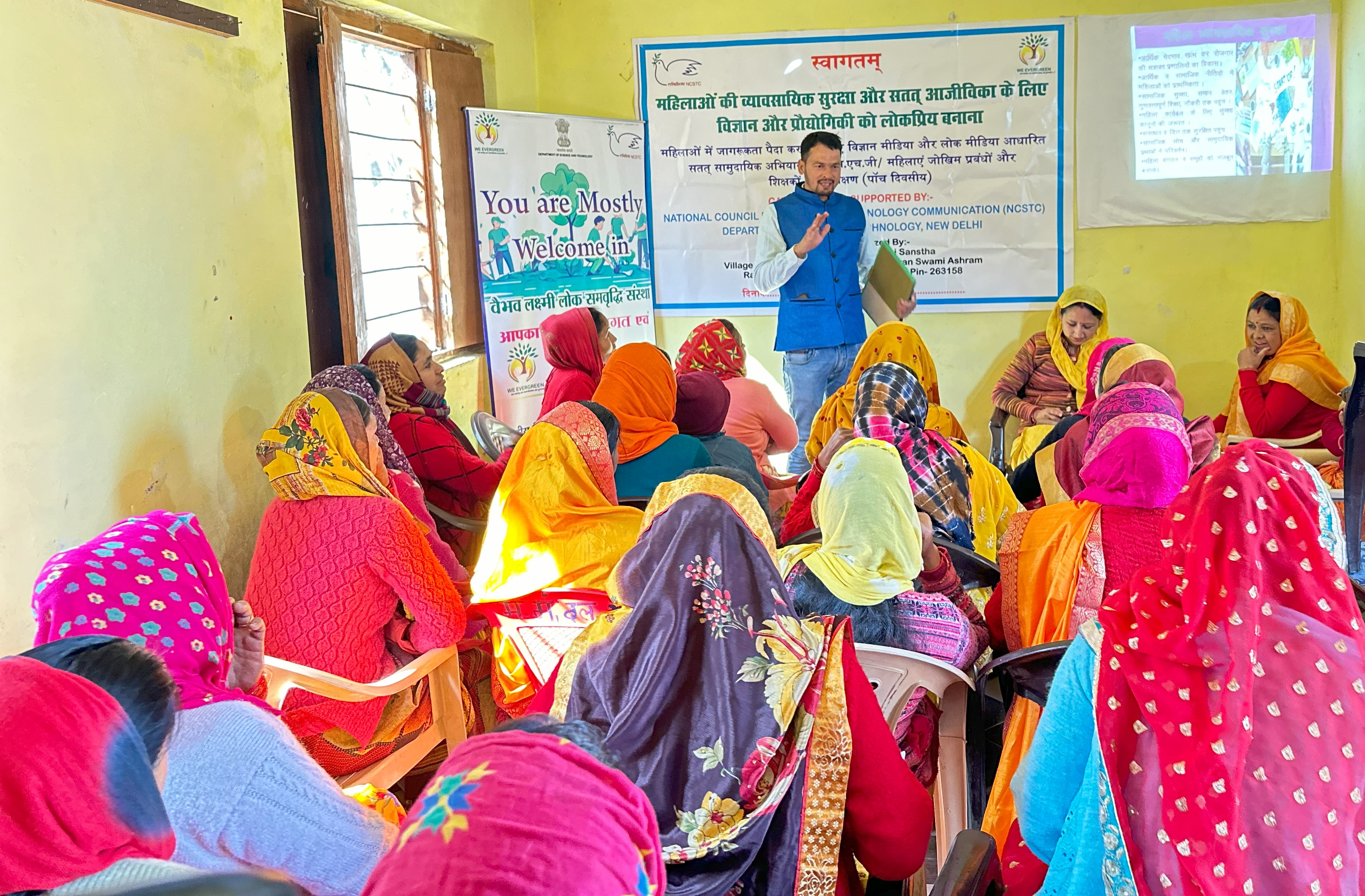 A trainer speaks to women in a classroom beside a programme banner and a projected presentation.